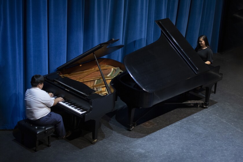 A boy and a girl play the piano on two separate pianos facing each other. The boy is wearing blue pants and a short sleeve button up shirt. The girls is wearing a black top.