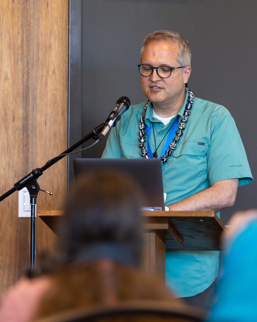 Author David Pulsipher wears a kukui nut lei and a teal blue shirt as he speaks at a microphone at the conference.