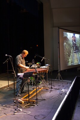 portrait shot of music professor Darren Duerden on stage with his xylophone and to his left a screen with a silent film playing