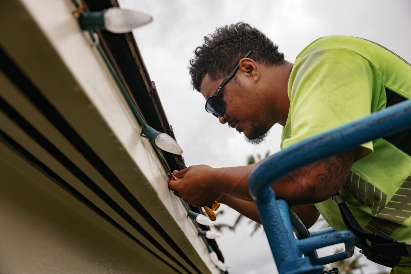 A worker wearing a bright green t shirt and black sunglasses attaches lights to the edge of a BYUH building.