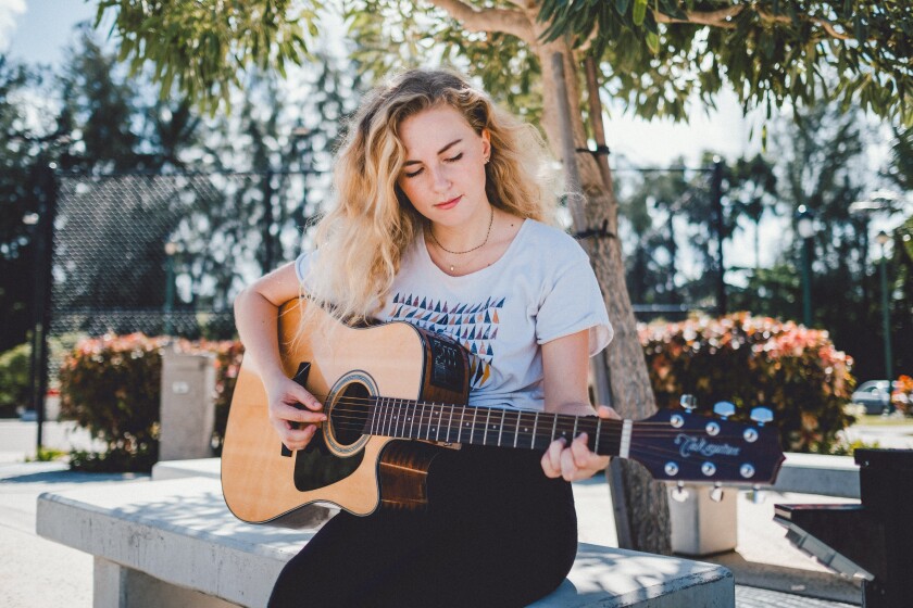 Danielle Knox sits on a bench in the courtyard on a sunny day and plays her guitar.
