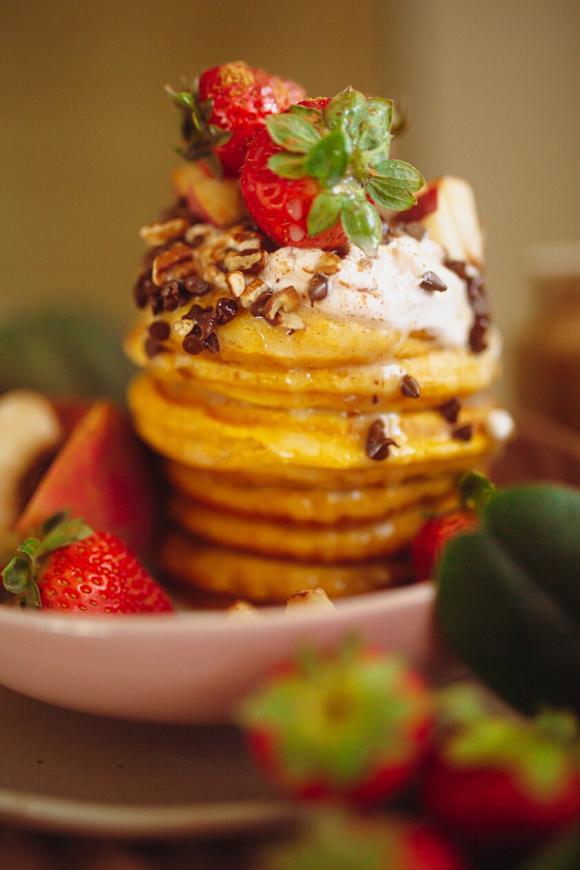 A stack of pancakes on a pink plate topped with whole strawberries, chocolate chips, nuts and whipped cream. Extra toppings are visible in the foreground.