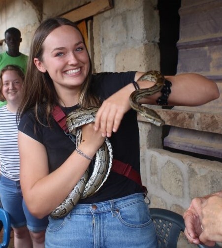A girl with light-brown hair wearing a black shirt and jeans holds a green snake in her arms.