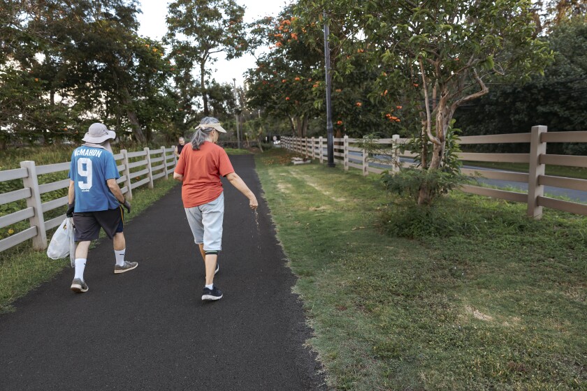 John (wearing a blue shirt with the number nine on it) holding a plastic bag walking next to Rhonda Bell (wearing a red shirt and grey shorts) who is dropping bird seeds on a walking path.