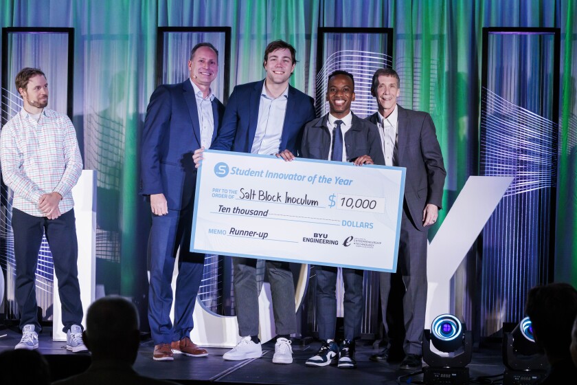 Four men stand in the shot. The man on the far left is not part of the group, he is a presenter. The three young men hold a check.