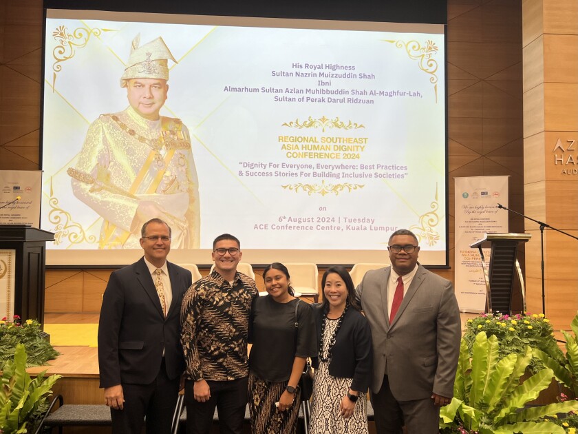 Two male professors, one female professor, one male student, and one female student pose in front of a presentation that says, "Regional Southeast Asia Human Dignity Conference 2024."