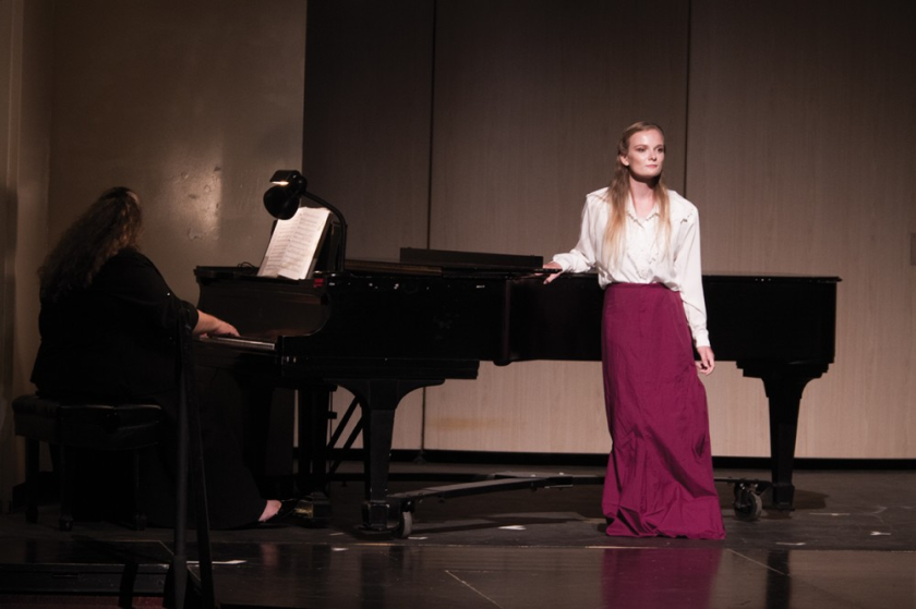 A woman in a dress standing next to a piano. Another woman sits at the piano.