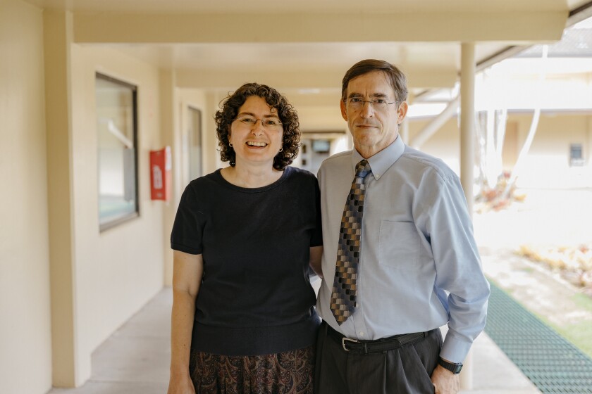 Jennifer Lane smiling, wearing a dark blue shirt with a yellow, blue and purple patterned skirt, standing next to Keith Lane, wearing grey suit pants, a blue button-up shirt and grey, blue and grey tie with a hallway behind them.