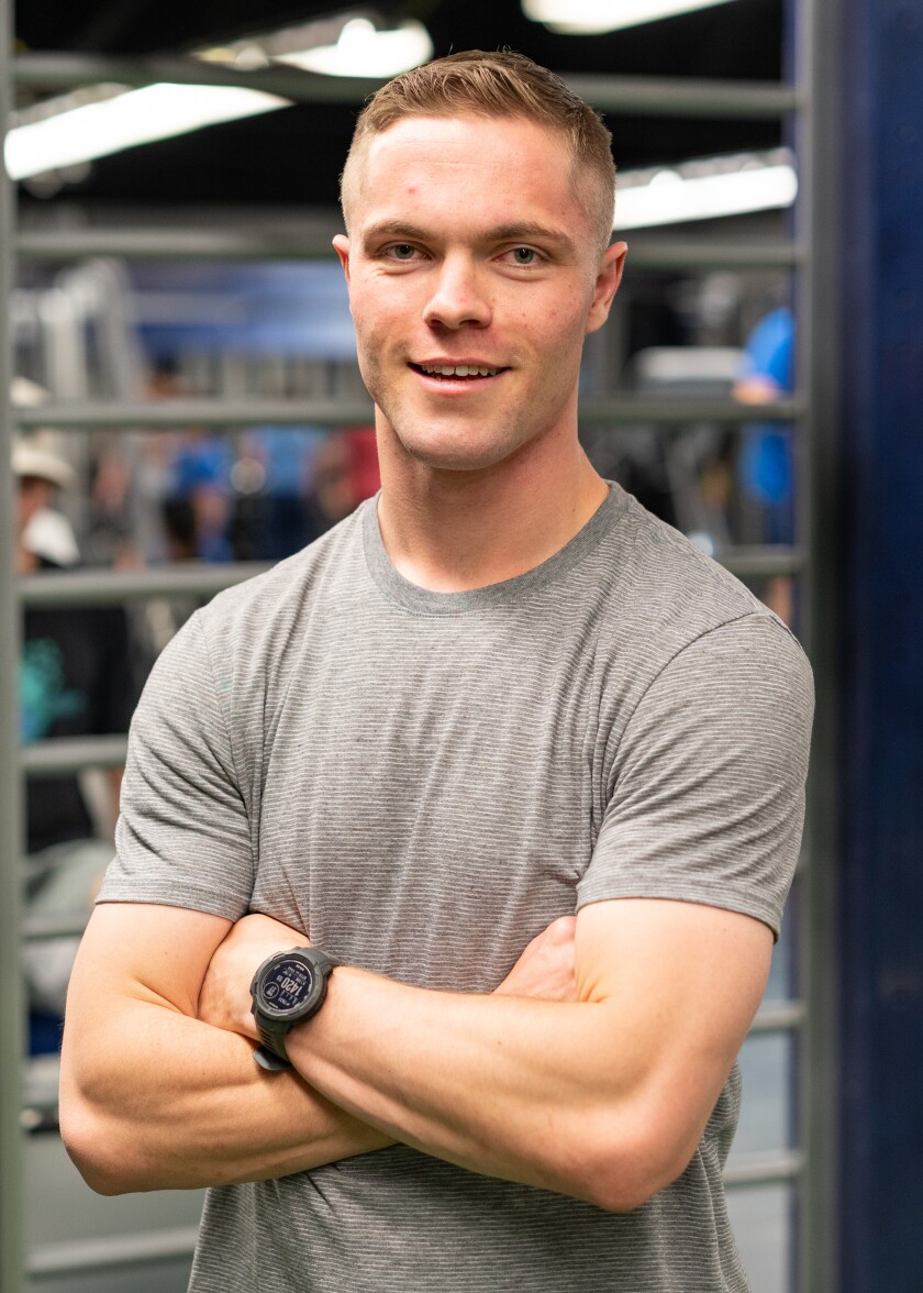 Portrait of a male college student with short hair in a gray t-shirt standing with his arms crossed. There is gym equipment in the background.
