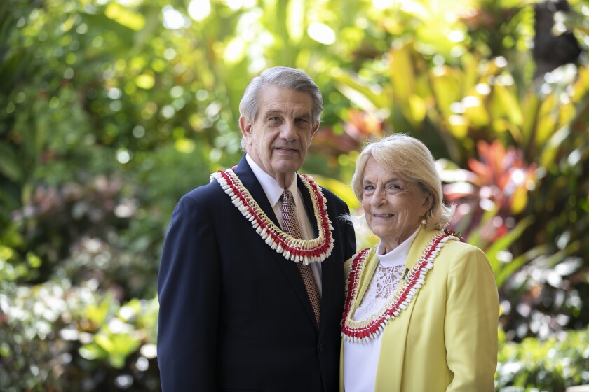 President Hallstrom wearing a dark blue suit and red and white poka dotted tie smiling next to Sister Hallstrom, wearing a white blouse and yellow jacket, both wearing red and white leis with greenery behind them.