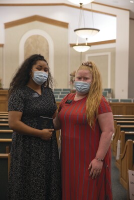 portrait shot of two sister missionaries standing in the Stake Center chapel at BYUH wearing masks and one of them holding a Book of Mormon