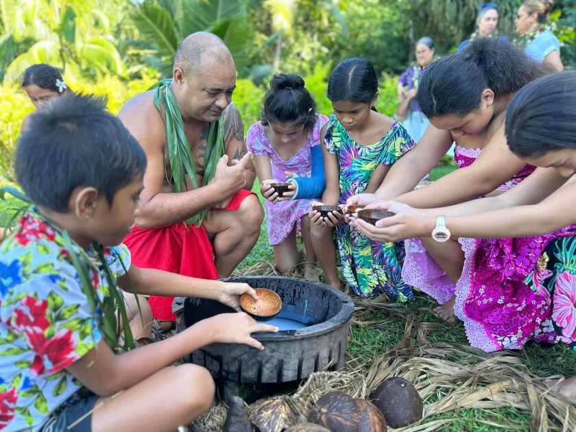 A group of young children and a Polynesian man in traditional clothing gather with coconut cups in their hands around a pot on the ground.