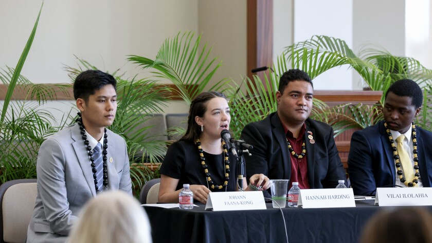Three men and one woman dressed professionally sit at a table speaking to a crowd.