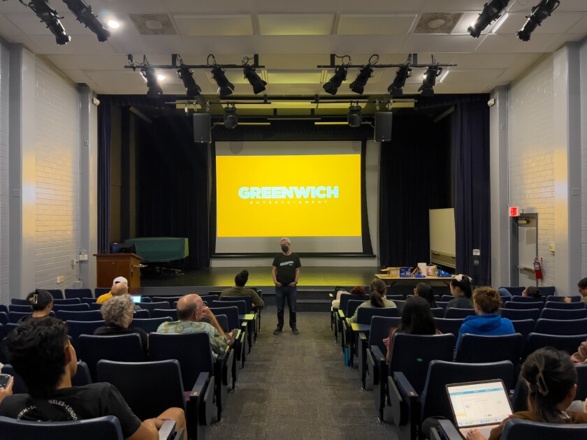 A man standing in the center of a room with a screen displayed on the back and people sitting facing him.