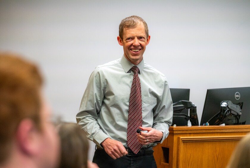 A male professor wearing a grey shirt and a red tie stands in front of a lecture room smiling.