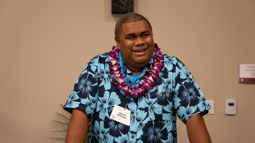 Associate Professor David Whippy wears a blue flowered aloha shirt and a purple and white orchid lei as he stands and speaks during the conference.