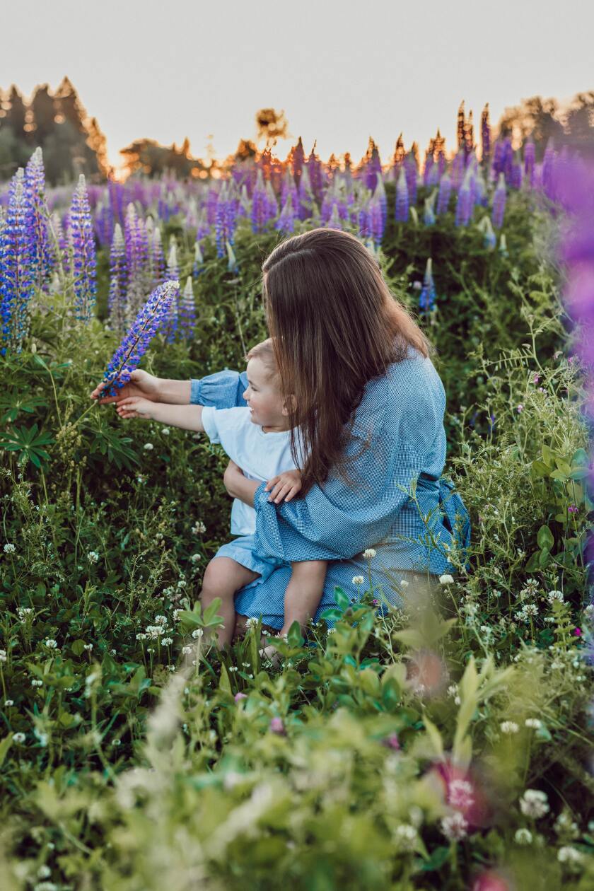 A woman in a blue print dress holds a toddler in a field of flowers as they look at a flower growing next to them