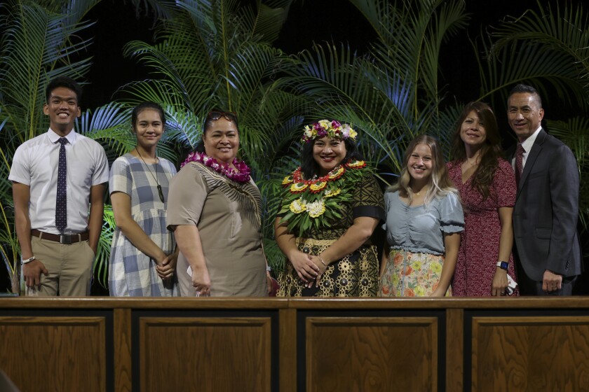 Men and women stand in button-up shirts and dresses side-by-side with a wooden divider in front of them and plants behind them.