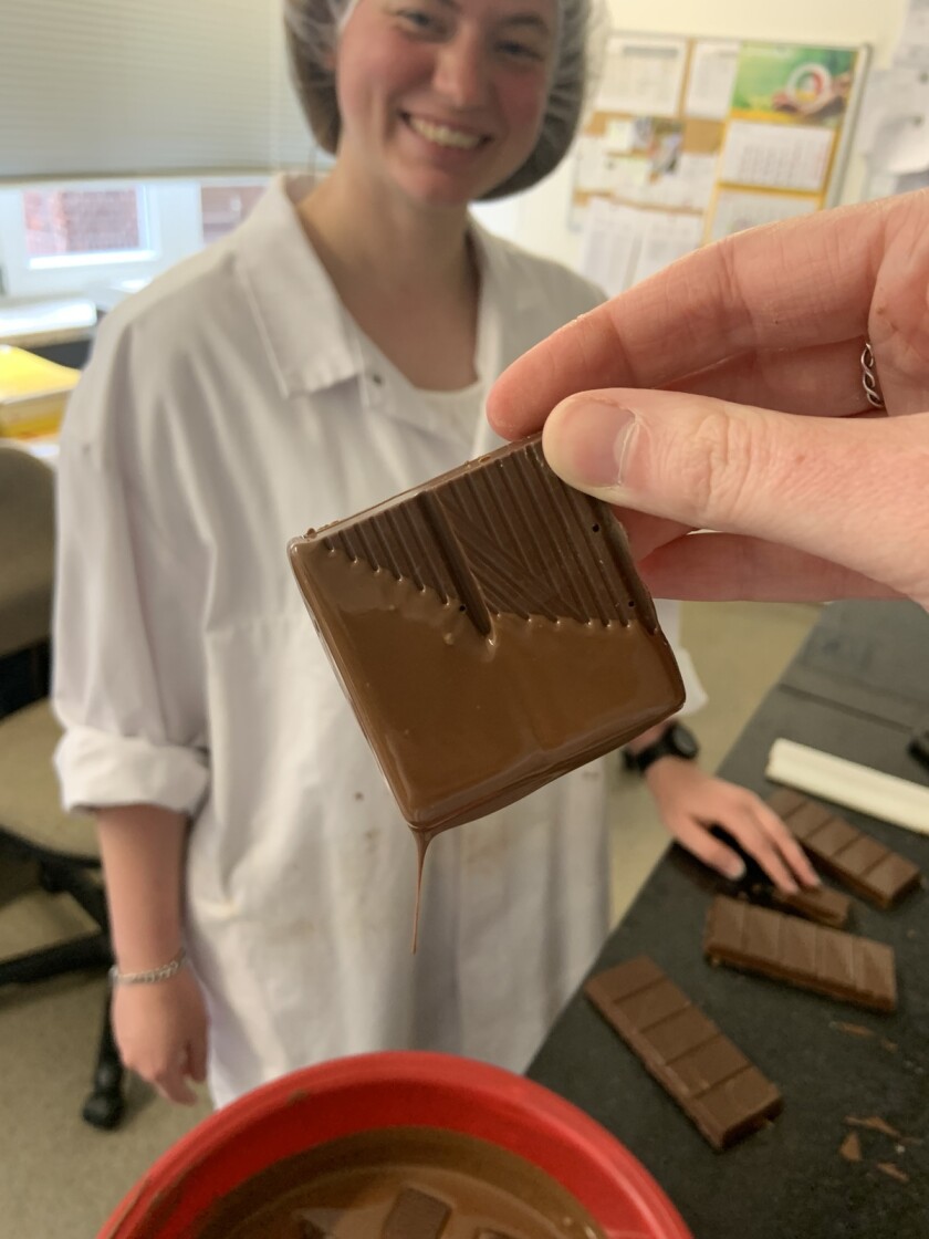 Erin's hand holding a chocolate square dipped in liquid chocolate. Behind the chocolate, Janece Nufer stands smiling, wearing a white lab coat and hair net. There are more chocolate section on a nearby table.