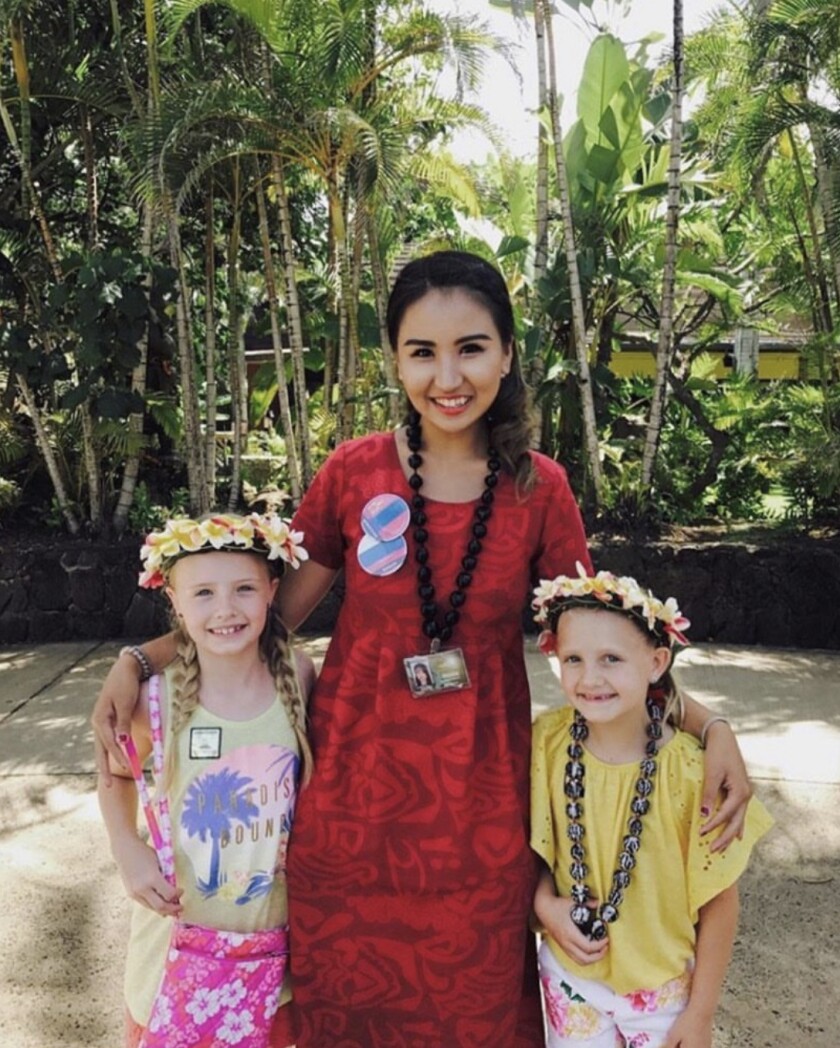 Nomin Khosbayar stands in red dress as a tour guide at the Polynesian Cultural Center with two girls wearing leis on their heads.