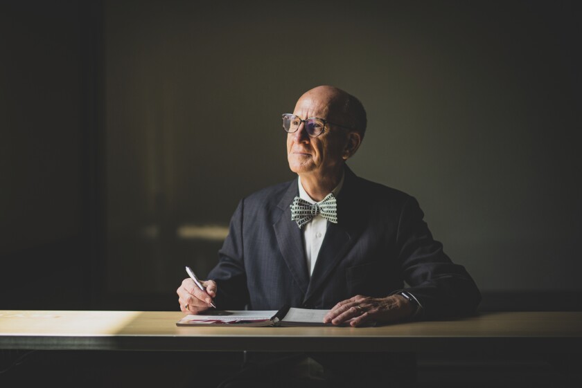 Dr. Neil Anderson looks to his left sitting down writing on a notebook, wearing a black tie with a black-spotted bowtie and glasses reflected on the light.