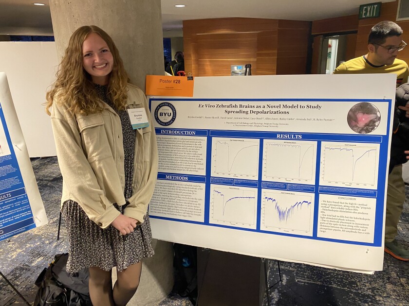 A girl in a beige jacket and black dress smiles in front of a presentation board.
