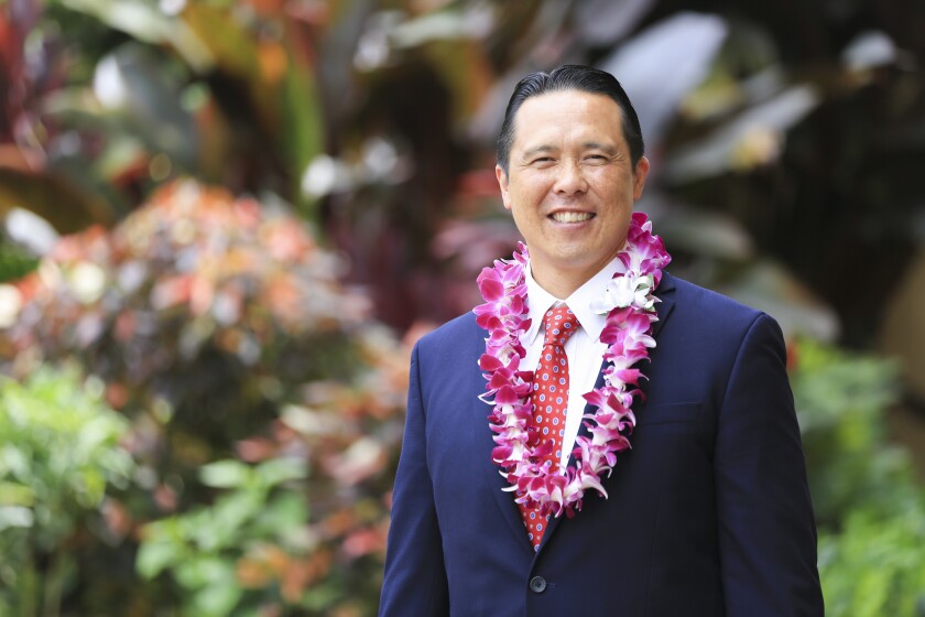 Taran K. Chun smiles wearing a black suit and red tie with a pink/purple flower lei and greenery in the background.
