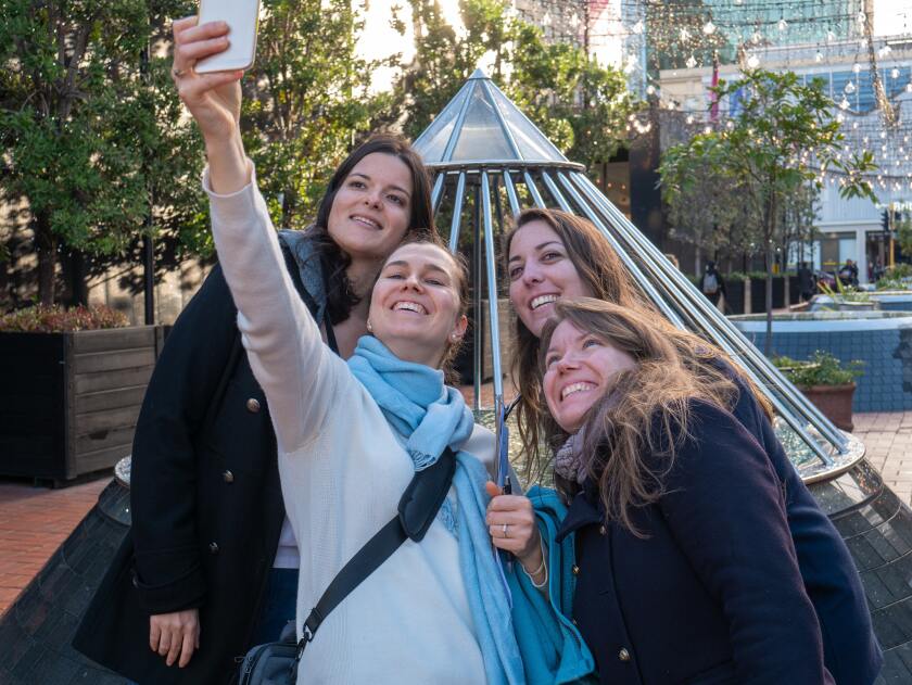 landscape photo of four women taking a selfie while wearing sweaters and scarves in front of a triangular structure somewhere in a city with trees and lights in the background