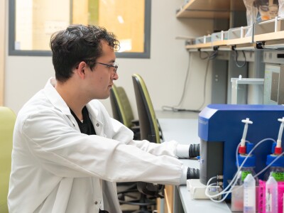 Israel Aleman, a young man with curly, black hair and glasses, processes data at a computer. He is wearing a white lab coat and black, latex gloves.
