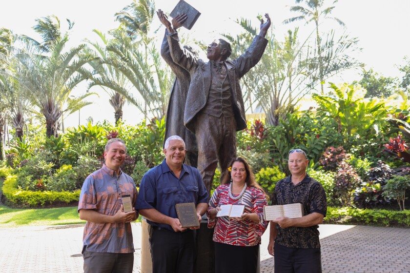 Robert Lono Ikuwa, Keali‘i Haverly, Alohalani Housman and Kamoa‘e Walk hold physical and digital Hawaiian scriptures in their hand with a statue of Jonathan Napela and George Q. Cannon behind them.