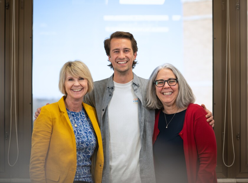 A young adult light-skinned male with short brown hair smiles, wearing a white shirt with a grey button up shirt over it, with his arms around two smiling light-skinned women, one with short blonde hair and a yellow blazer and blue blouse and the other with black glasses, shoulder-length grey hair and a red cardigan and black shirt.