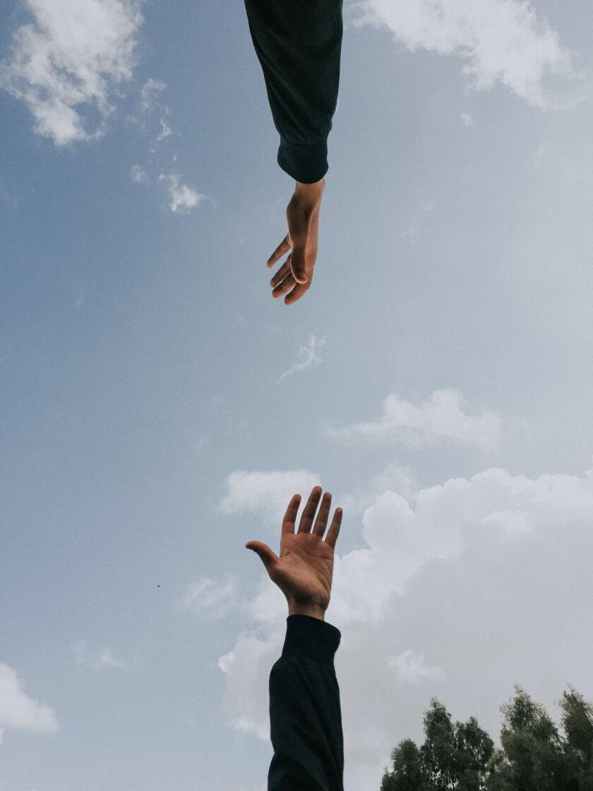 Two arms, both with black long-sleeved shirts on, reaching out to each other. Behind the hands is a blue sky and there are green trees in the right corner.
